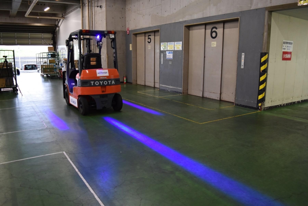 倉庫内を走行するフォークリフトの床面に青色のLEDラインが投影され、周囲の作業者に接近を知らせて接触事故を防止している様子を示す